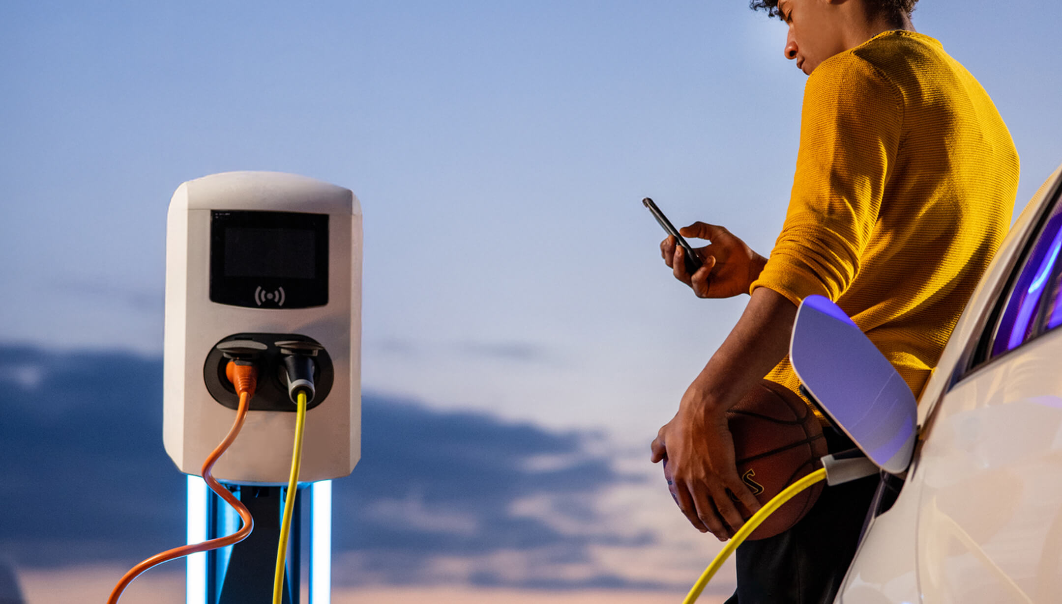 A boy with a cellphone and a basketball between an electric pole and a car charging