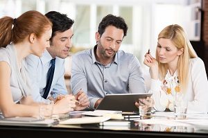 Confident businessmen and businesswomen sitting and working on tablet during a meeting. Business people sitting around the desk in a meeting and watching digital tablet. Portrait of happy colleagues looking at laptop screen in office.